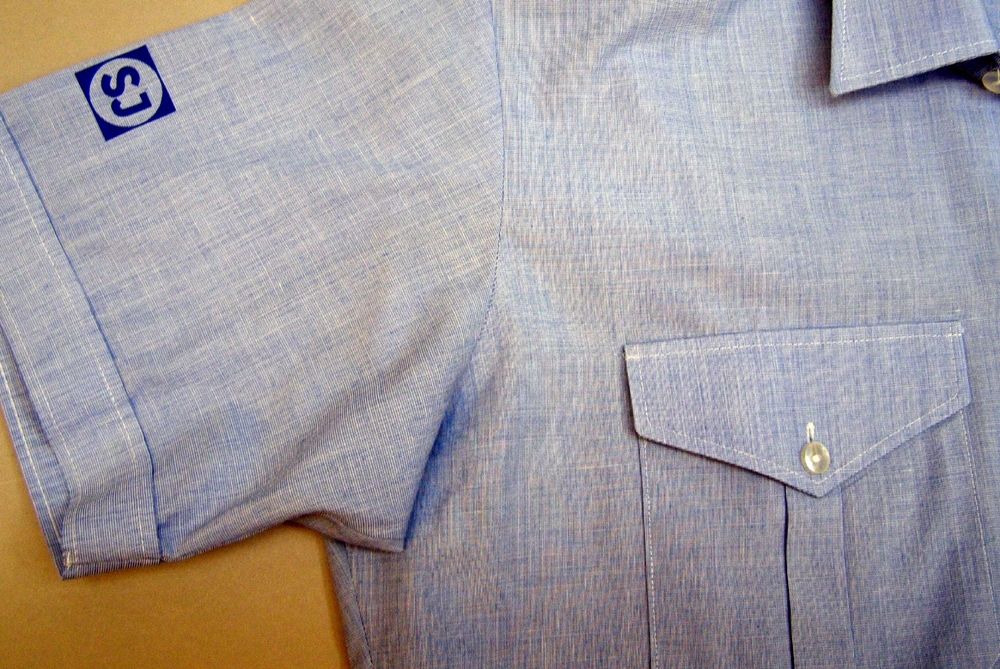 Kortärmad skjorta av tunnt polyester-bomullstyg, ljusblå/vit-melerad med ok, krage och två bröstfickor med lock och knäppning. Skjortan knäpps fram med med små knappar av vit plast. Märkning/tryck i mörkt blått på höger ärm ovantill: "SJ" i fyrkant i cirkel. Tillverkningsmärkning på okets insida: "Made in Norway Selje skjortefabrikk".