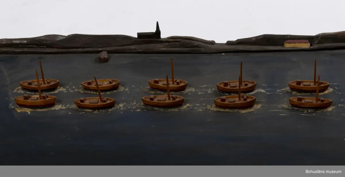 Rektangulär träplatta med fjorton båtar, två och två, i en rad.
I fonden är det en siluett av ett kustlandskap med berg, ett hus och en kyrka.
Text framtill på plattan: "SKARPSILLSFLOTTAN PÅ VÄG GENOM GULLMARSFJORDEN".
En större båt saknas. Master saknas på vissa av båtarna.

Ur handskrivna katalogen 1957-1958:
Skarpsillfångstflottan
Modeller på platta. Plattans mått: 116 x 42. Föremålen hela.
Från kapten Olssons saml., Fiskebäckskil.

För ytterligare information om förvärvet, se UM005087.