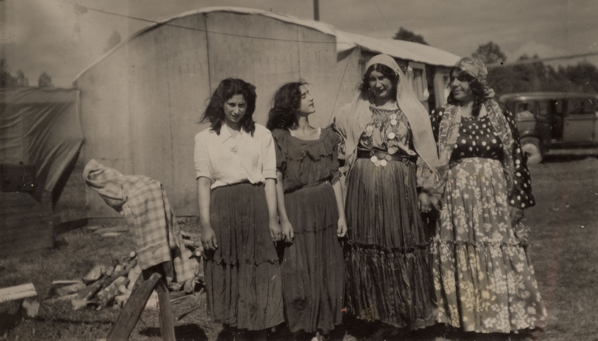 Fyra romska kvinnor framför ett tält och en bil. De två äldre kvinnorna till höger bär "dikhlo", en huvudduk som markerar att de är gifta. En av dem bär även ett halsband av mynt. Fotografiet är troligen taget i Sandviken 1947.
