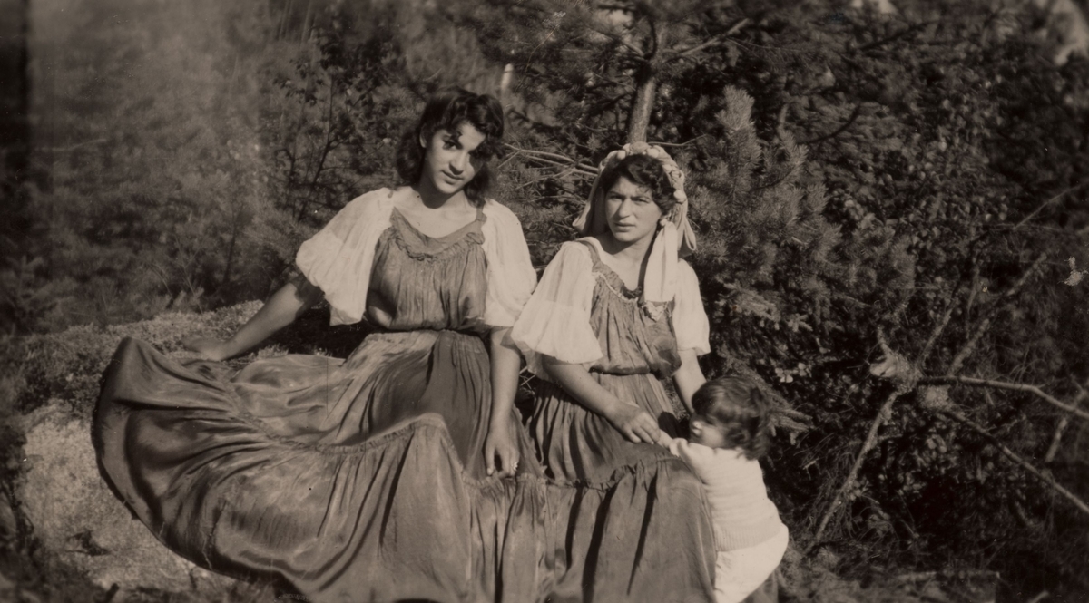 Två romska kvinnor sitter på en bergsknalle en sommardag i Hofors 1950. Ett barn är på väg att klättra upp i den ena kvinnans knä. Bakom dem syns barrträd.