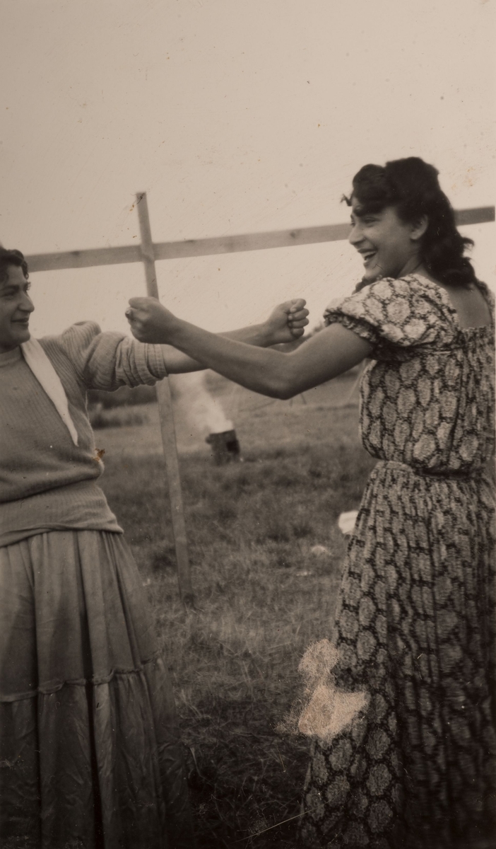 Två romska kvinnor hytter med nävarna mot varandra och skrattar. I bakgrunden syns en rykande tunna på en äng. Fotografiet taget i Sandviken i juli 1950.