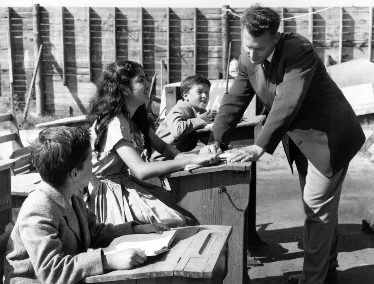 Skolundervisning en sommardag på Limhamnsfältet i Malmö 1955. De tre eleverna sitter vid inlånade skolbänkar i det gröna och lyssnar till läraren.