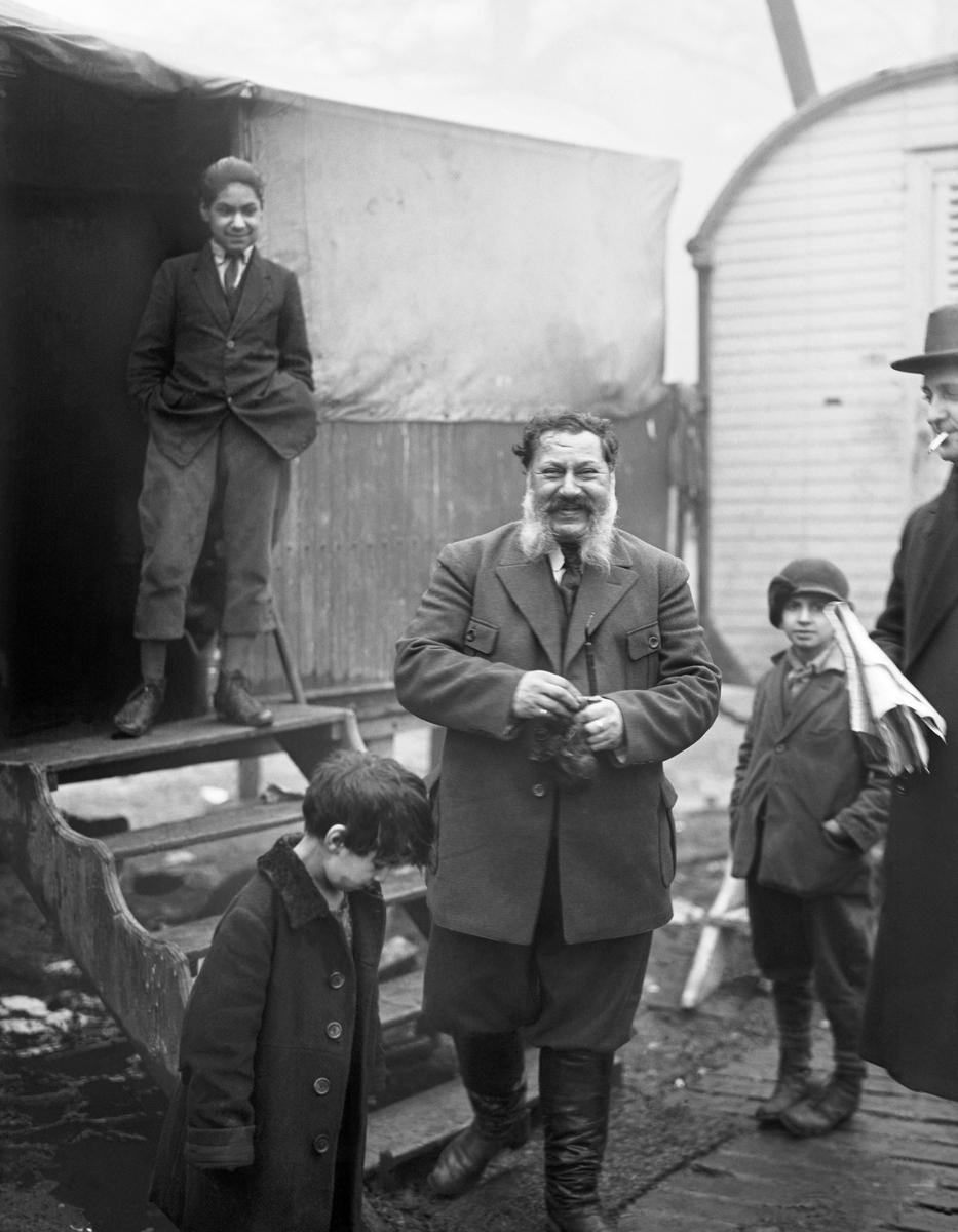 En romsk lägerplats med bostadsvagnar och tält. På bilden syns en äldre man med stort skägg samt tre barn i olika åldrar. I förgrunden står ytterligare en man i kappa, hatt och håller i en dagstidning.