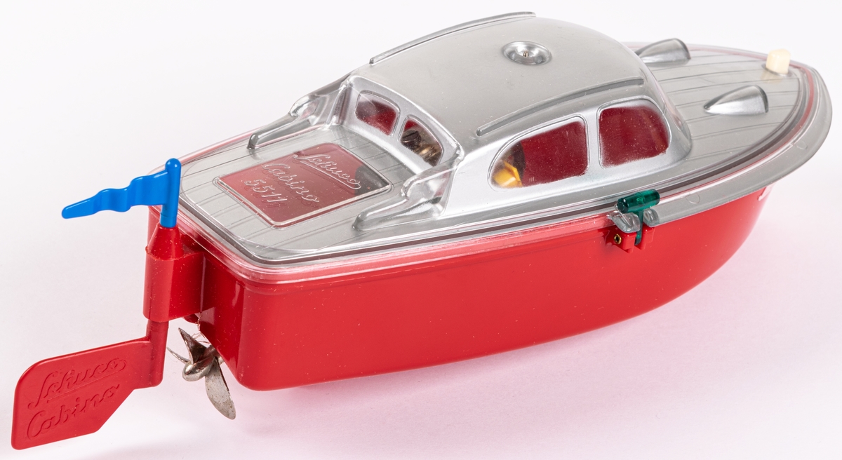 Leksaksbåt av gjuten hårdplast. Rött skrov och grått däckparti. Drivs med batteri. Ett batteri finns kvar. Båten ligger i orginalförpackning med bruksanvisning på engelska. Modell Cabino 5511. Tillverkad av Schuco i Tyskland.