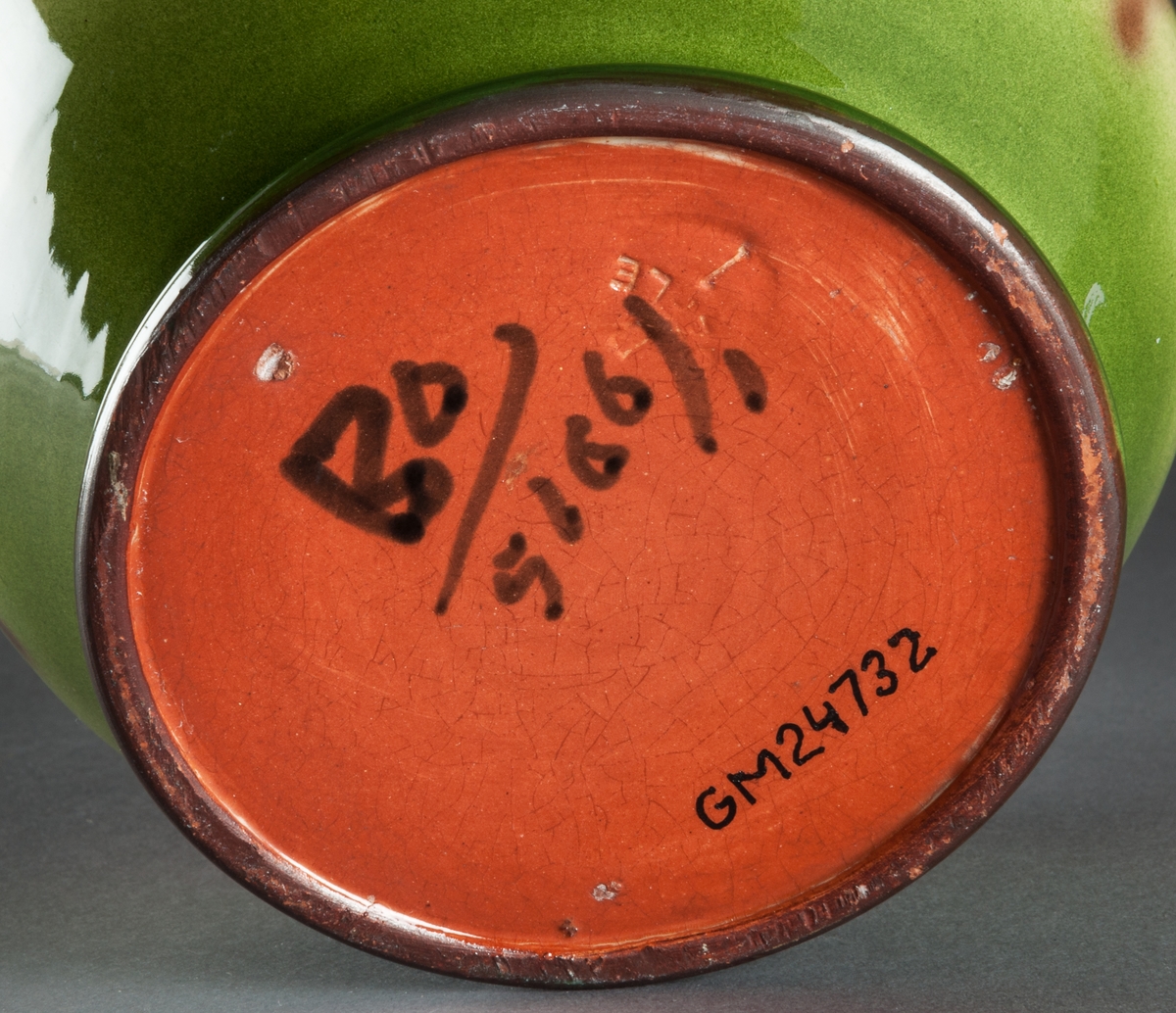 Kruka av keramik. Handdrejad. Bukig med låg utsvängd profilerad mynningskant. Glaserad med flammiga gröna färger och blommor i brunt. Färgmärkt under "Bo/5116/". Tryckstämpel "B Gefle 1". Modell 5160 anger att Gabriel Burmeister har formgivit ytterkrukan. Möjligen är den producerad och glaserad efter hans anställningstid (1922-24).