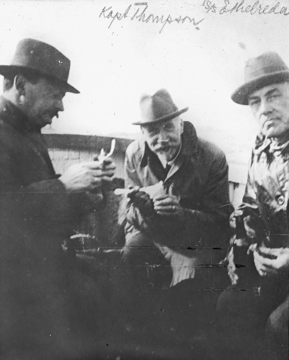 Nærbilde av tre menn i frakk og hatt ombord på et fartøy. De spiser mat. Merket Kapt. Thompson og S/S ETHELREDA.