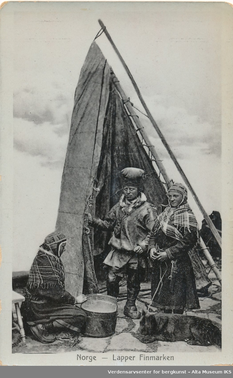 En samisk mann står sammen med en samisk kvinne. Hun har en hund i bånd som ligger på bakken og hviler. En annen kvinne sitter på huk foran dem med en eske/kiste.