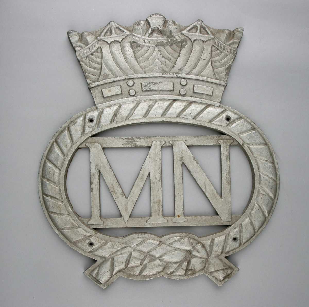 Ovalt emblem med krone. I senter bokstavene MN. Emblem ser ut som tau med båtsmannsknop.