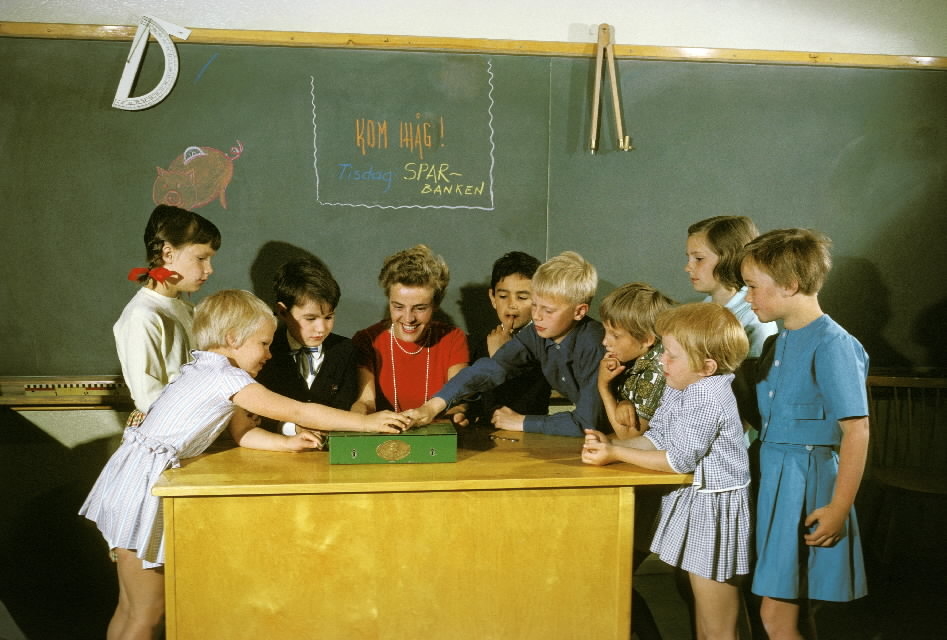 Seriebild L 4 a, alt 1. Skolsparsituation i klassrum i
Bergshamraskolan, Solna. Barnen lägger sina pengar i skolsparbössan
under lärarinnans överinseende. På svarta tavlan står uppmaningen.
"Kom ihåg! Sparbanken"