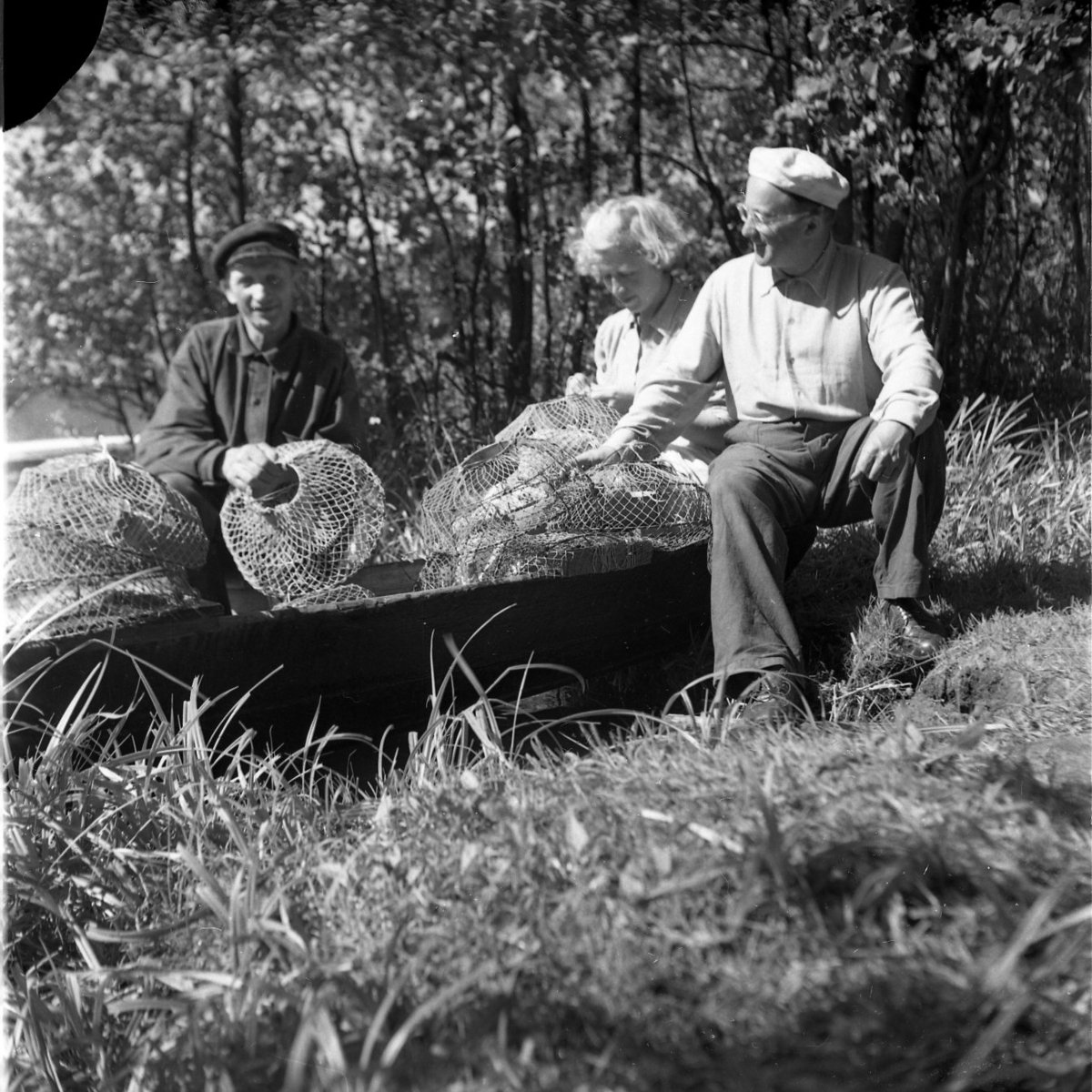 Två män och en kvinna sitter möjligen intill en eka med kräftmjärdar. Det är kräftpremiär. Mannen till vänster har keps och heter Einar Karlsson. Till höger sitter herr och fru Eckerberg.