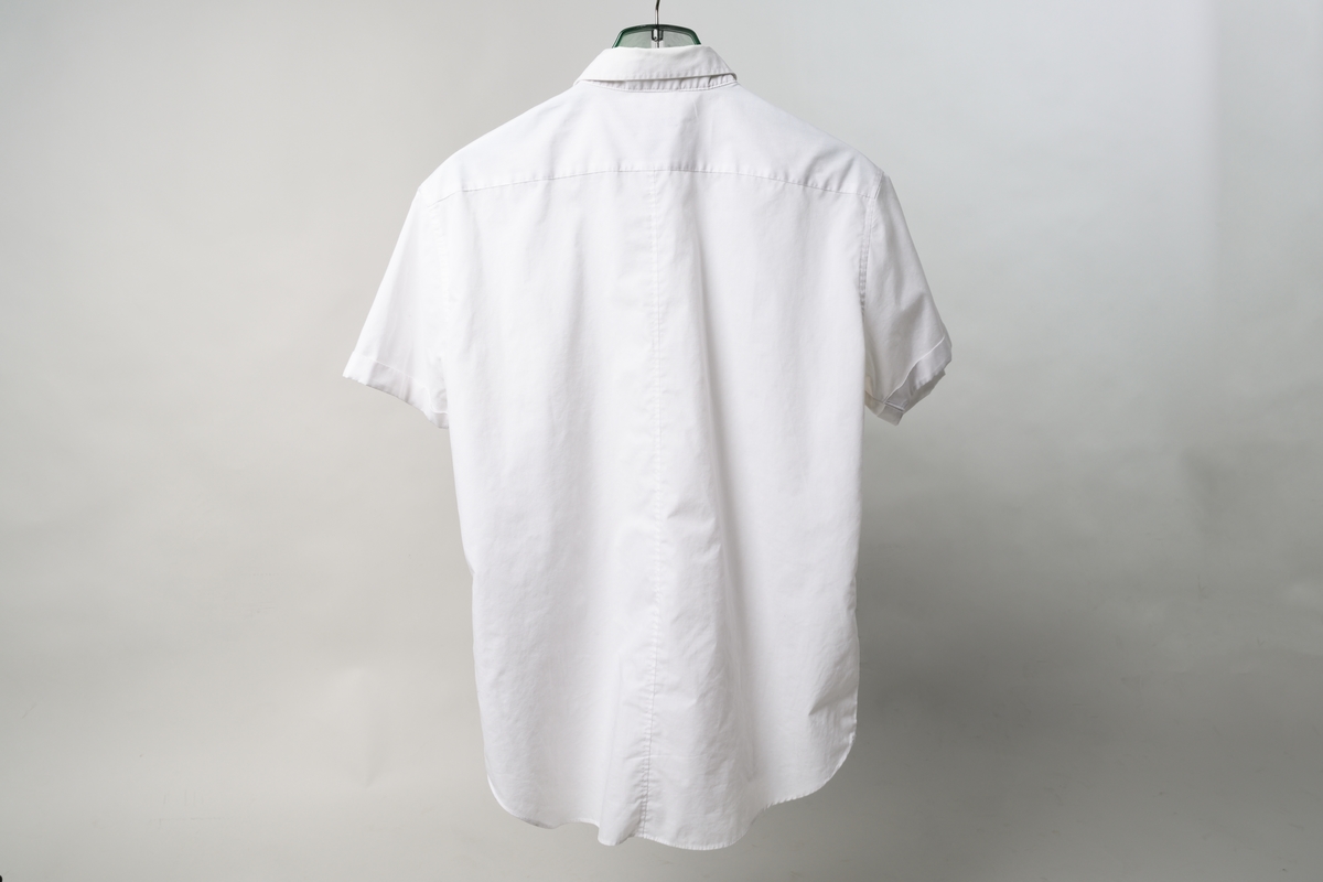 Herrskjorta av vitt tyg (65% polyester, 35% bomull), med korta ärmar med slag, krage och baktill ok. Knäppning fram med sju plastknappar. På axlarna klaffar med knäppning med plastknapp. Storlek 40/42.