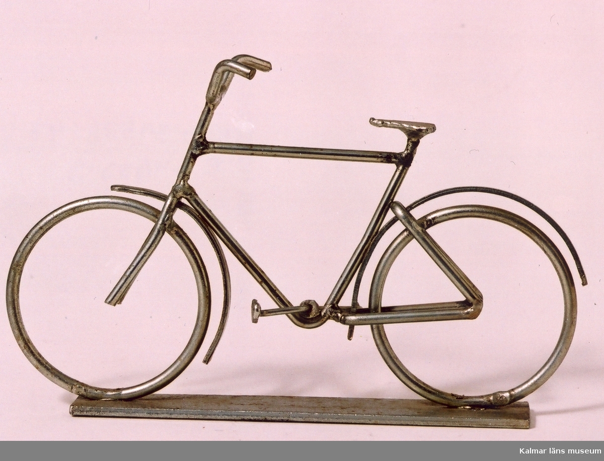 KLM 39586:47 Cykel av metalltråd, omålad. Tillverkad i konstsmide, svetsad. Herrcykel med vanligt styre. Monterad på en metallplatta. Prydnadsföremål. Tillverkad av Yngve Axtelius som var verksam som trådslöjdare.