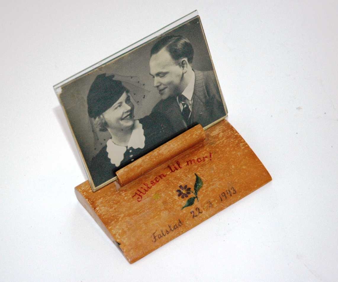 Bildeholder i tre med påmalt blomstermotiv og tekst, datert Falstad 22.03.1943. I bildeholderen er det satt et foto av Leif Hallesby og hans kone Else. Bildet står mellom to glass.
