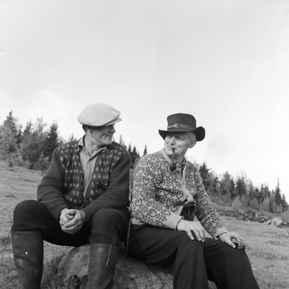 Dagfinn Grønosets foto av Laurits Engen og Ole P. Bjørnstad. To menn som sitter på en stein og ser på hverandre, den ene røyker pipe.