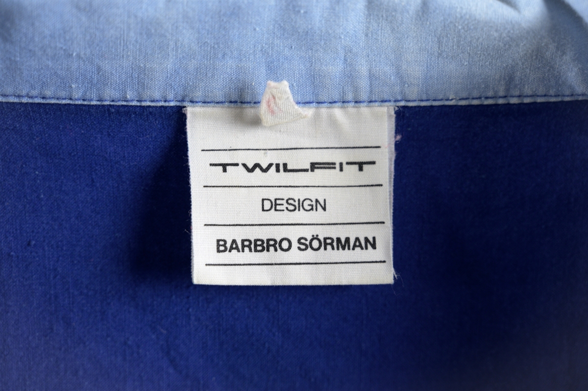 Skjorta designad av Barbro Sörman för Twilfit. Skjortan är kornblå med detaljer på ärmar, fickor och krage i ljusblått och mintgrön 100% bomull. Vid modell som dras över huvudet, krage och slag samt att det är sprund på båda sidor, två stora utanpåliggande fickor nedtill, dessa har en kant av ljusblått tyg. Gröna klaffar och ljusblå kilar vid ärmen, mintgrön bård vid ärmslut. Baktill sitter en grön rektangulär detalj som ev. sytts dit i efterhand för hand och den täcker ett litet hål. Skjortan har några fläckar på framsidan samt att den troligtvis är något blekt.