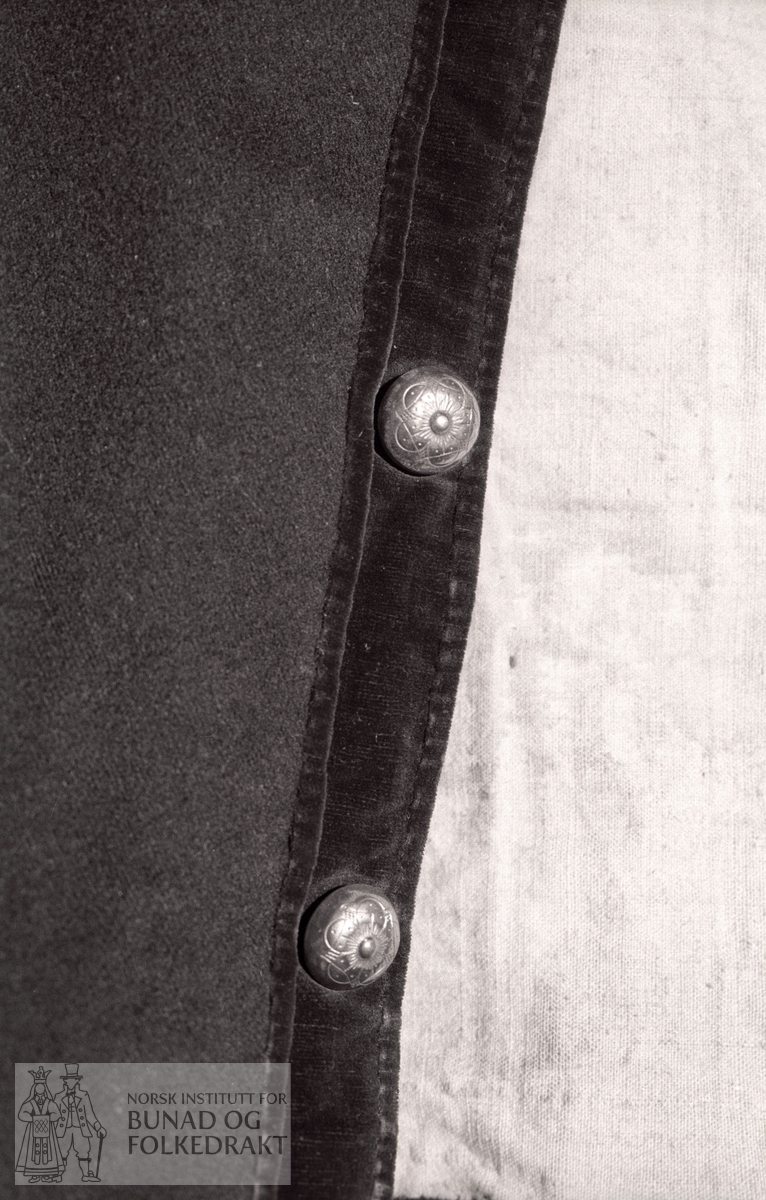 Svart klede, med brun fløyel. 6 kuva sølvknappar framme. 1 flat knapp ved ermesplitt. H. rygg: 36,5 cm. H. framkant: 43 cm. Br. ved sømmer bak 1 cm (i livet). H. krage: 5,4 cm.