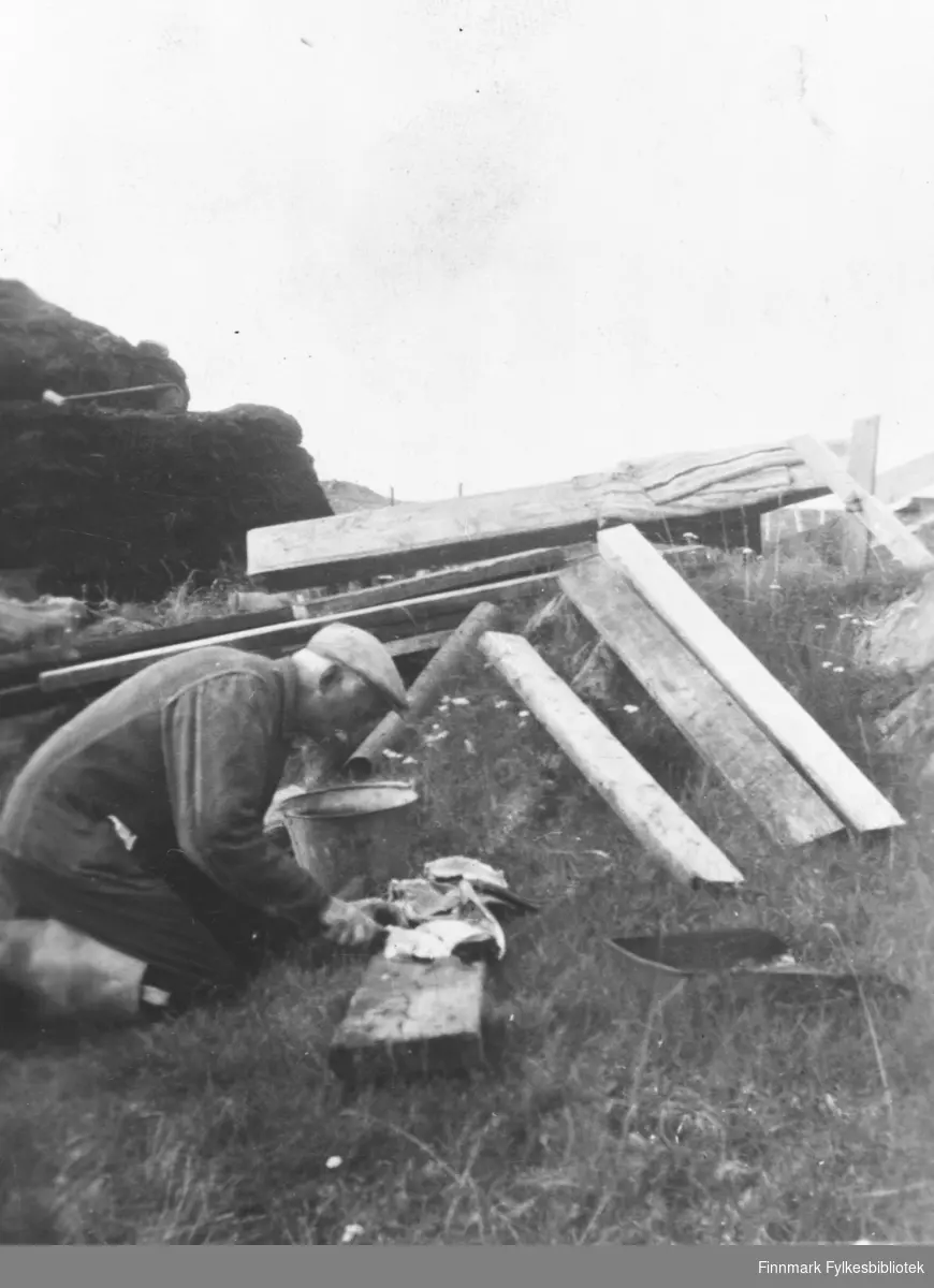 Georg Adriansen sitter på kne og sløyer fisk på ei fjøl. Bak ham ligger det tømmer og plank. Bildet er tatt etter brannen i 1945 i Gamvik. Folk var nødt til å klare seg med det de hadde. Georg ble tatt av tyskerne da de var på brenningstokt, og han satt på Grini til krigens slutt.