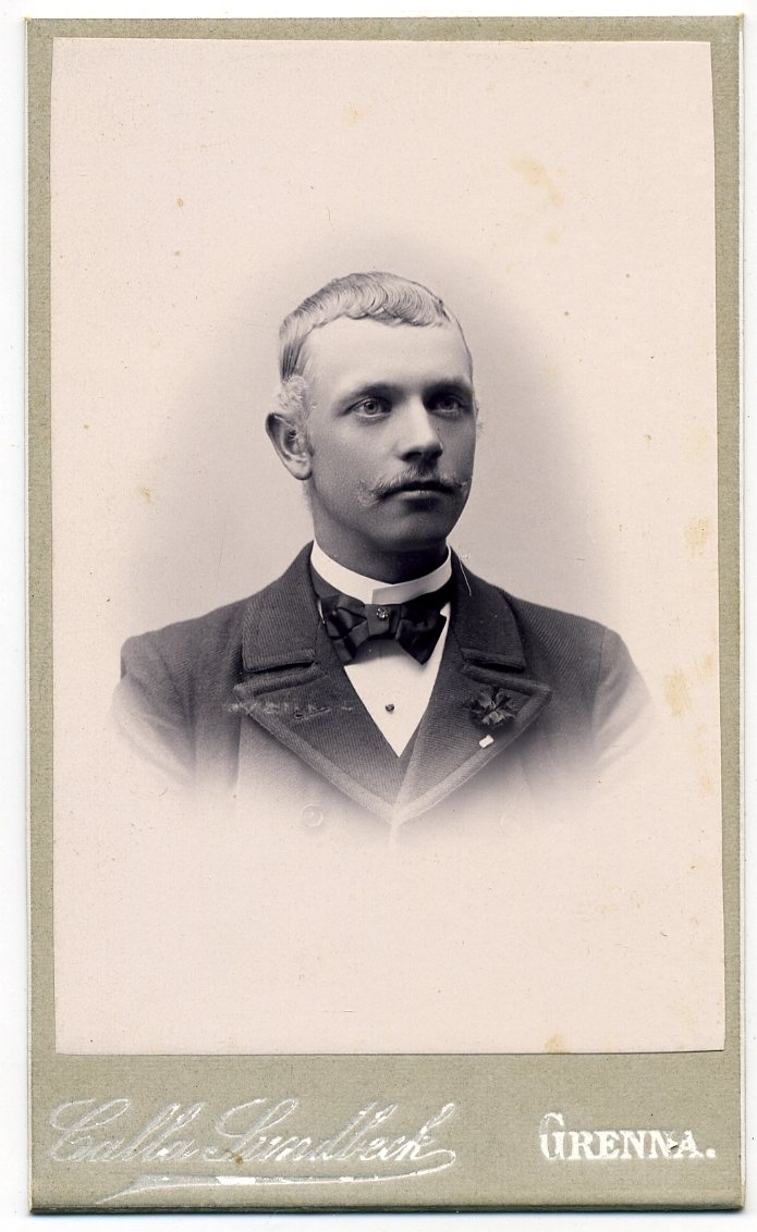 Kabinettsfotografi: porträtt okänd ung man med fluga. En liten brosch på flugan och en blomma i kavajuppslaget.