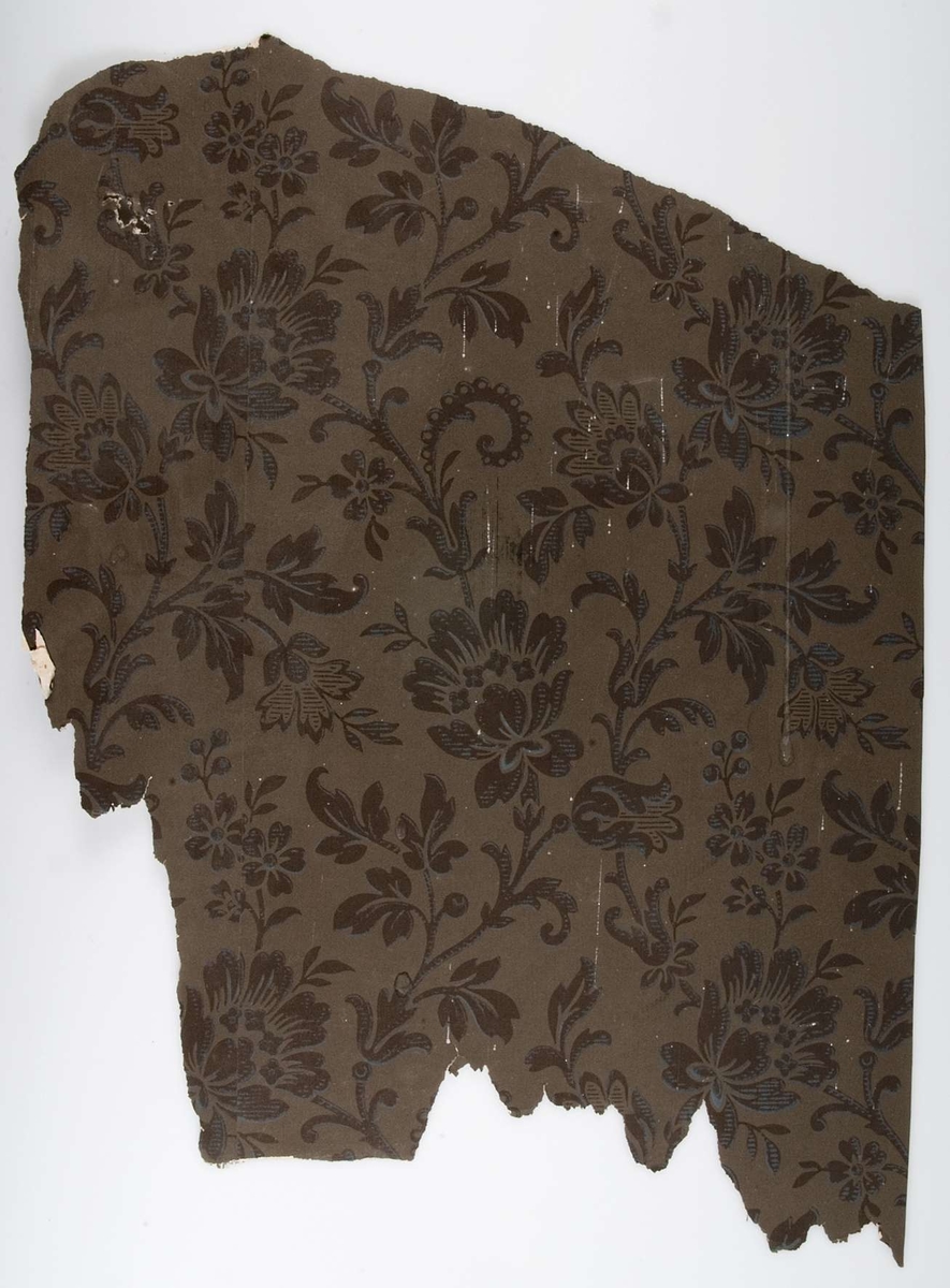 Ett fragment av tryckt tapet. Brun botten med växtmönster i brunt och blått. Tapeten dateras till 1887 (på tapetens baksida sitter tidningssidor från Upsala-Posten 2 mars 1887).