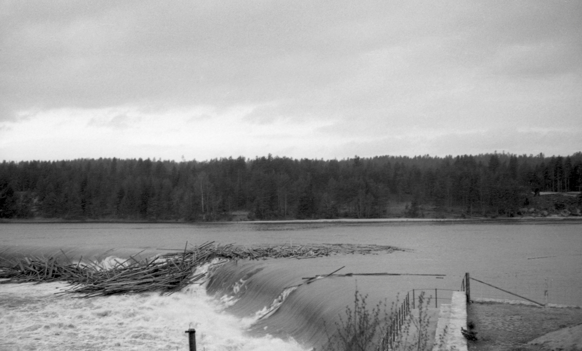 Tømmerfløting ved Skjefstadfossen i Glomma (i Heradsbygda i Elverum) våren 1941.  Fotografiet er tatt fra det høye og kraftige landkaret som forbandt den drøyt 300 meter lange, støpte damkonstruksjonen med vassdragets østre bredd. På dette tidspunktet rant det vann over både den østre delen av den buete damkrona, som var den laveste, og den midtre, som var en aning høyere.  På den midtre delen av dammen hadde det bygd seg opp en tømmervase, antakelig fordi stokkene falt fra dammen og ned i et parti der elvebotnen var grunn, steinete og ujamn. Det å løsne slike tømmervaser, mens vannføringa ennå var høy og mulighetene for videre fløting var gode, var farefullt arbeid for involverte mannskaper og en kostnadsdrivende faktor for Glomma fellesfløtingsforening. Følgelig er det grunn til å anta at fotografiet ble tatt for å dokumentere slike vanskeligheter overfor kraftutbyggerne, som hadde plikt til å holde andre aktører med hevd på bruk av vassdragets skadesløse.