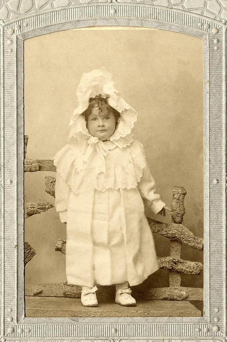 En liten flicka i lång vit kappa och stor vit hätta med brodyr. Hon står på en bänk framför en "gärdesgård". 
Helfigur, en face. Ateljéfoto.

Fotografens dotter.