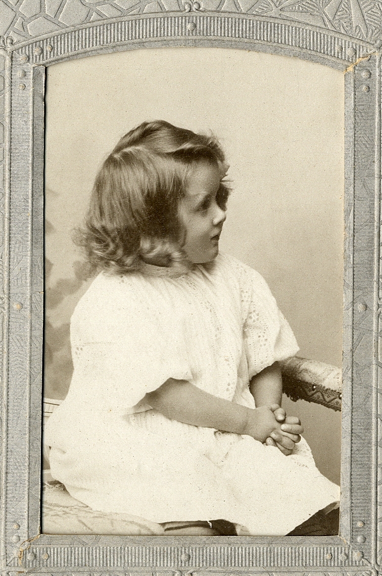 En liten flicka i ljus klänning, som sitter på en liten soffa. 
Knäbild, halvprofil. Ateljéfoto.
Jfr Alb14-094 

Fotografens dotter.