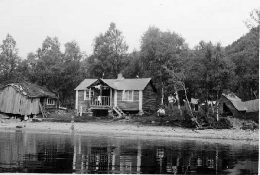 En hytte og et uthus ved et vann, et telt sees til høyre.