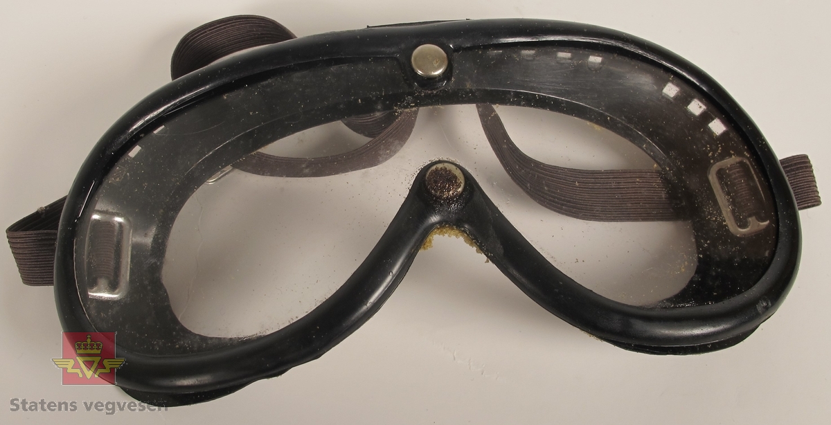 Briller i svart gummimateriale, med plastvindu som er festet med til brillen med to nagler. Det er rester etter skumgummi på den siden som er mot ansiktet. I gummien på siden og på toppen rundt brillen er det luftehull. Justerbart strikk festet til hver siden av brillen for justering til hode eller hjelm.
