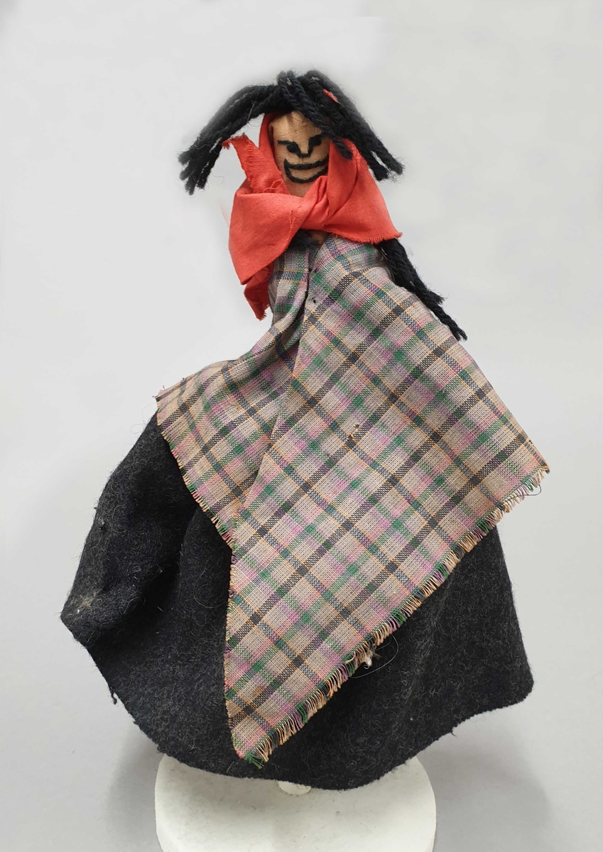 Dukke som fremstiller en trollkjerring, med svart hår av garn, rødt skaut, svart stakk og rutete sjal.