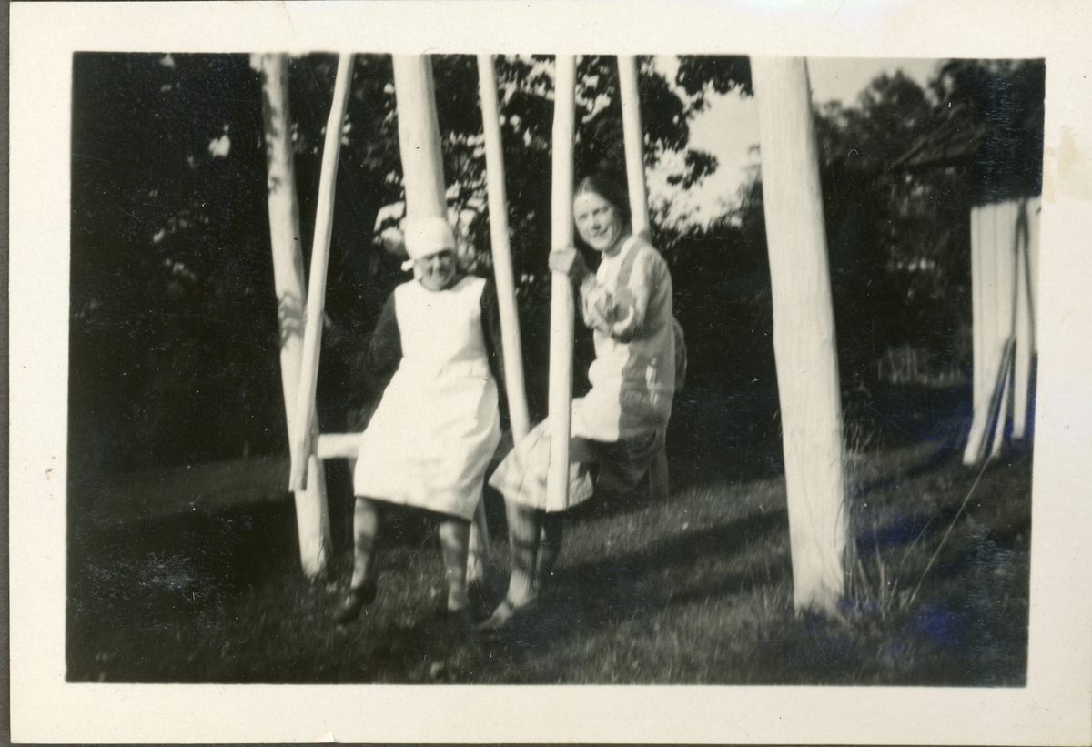 Elever fra husmorskolen på Skansgården, Kongsvinger, 1928. To kvinner, én i kjøkkenklær, i huskestativ i hagen.