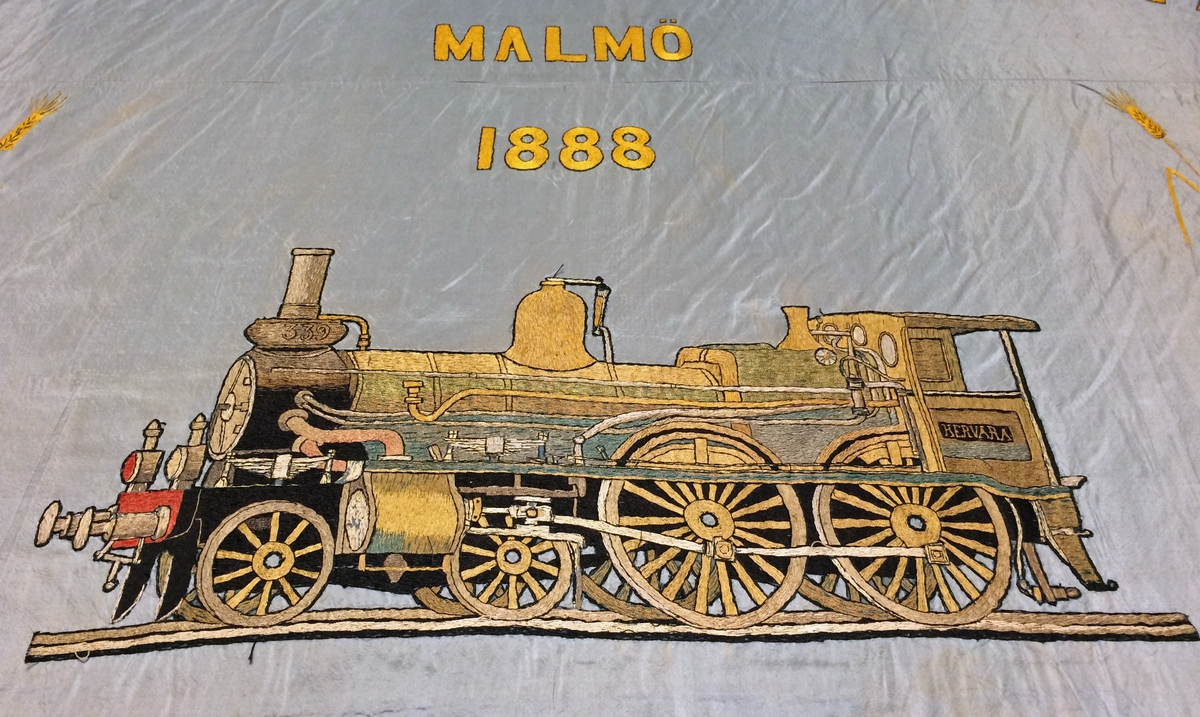 Rektangulär fackföreningsfana av blått broderat siden, för en järnvägsmannaförening i Malmö.
I mitten syns ångloket SJ Cb 339 Hervara, under detta ett bevingat hjul, en hjulaxel, samt ett arrangemang av verktyg och växtrankor.
Text i gult : "Personalen vid S.J. verkstad Malmö 1888 Arbete Enighet". Fanan är broderad i guld, svart och bruna nyanser med ett lok på mitten och eklöv i hörnen. Broderierna är utförda i stjälksöm och schattérsöm, vissa partier är även applicerade. Fanan har blågula fransar i kanterna.

Fanan är mycket blekt på den sidan som har legat uppåt i monter. Se sista bilden för jämförelse med undersidan.