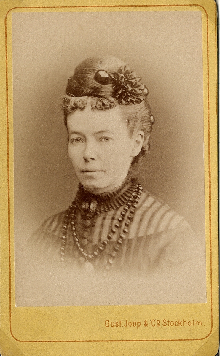 Porträttfoto av en kvinna i mörk klänning med dito tylljacka. Vid kragen syns en brosch och ett flerradigt mörkt halsband. 
Bröstbild, halvprofil. Ateljéfoto.