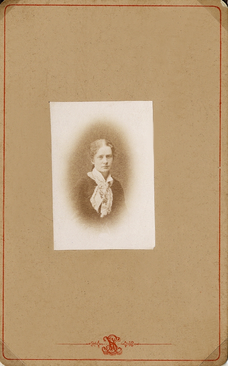 Porträttfoto av en ung kvinna i mörk klänning vit krage och knuten ljus sjal. Över sjalen syns en halskedja med medaljong. 
Midjebild, halvprofil. Ateljéfoto.