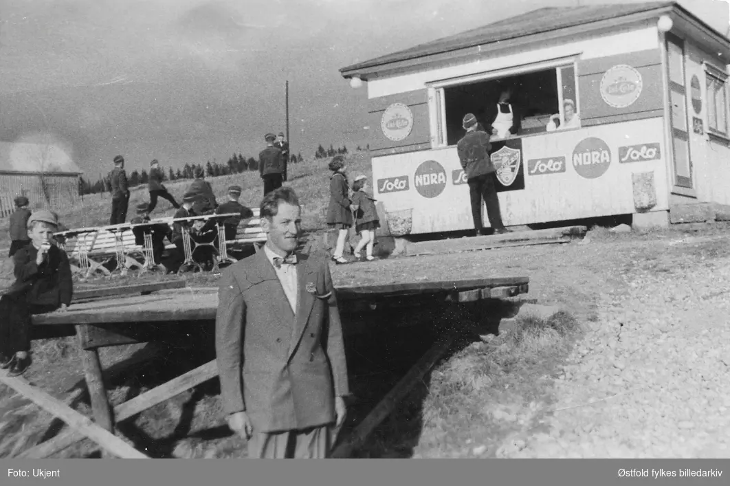 Anton Hauger ved pølsebua  til Solveig Grini "Berg gård", Spydeberg  1955. Barn i bakgrunnen spiser is og pølser, 17. mai?  Reklame for Solo og Nora. Musikkorpsmedlemmer, Spydeberg Guttemusikk.