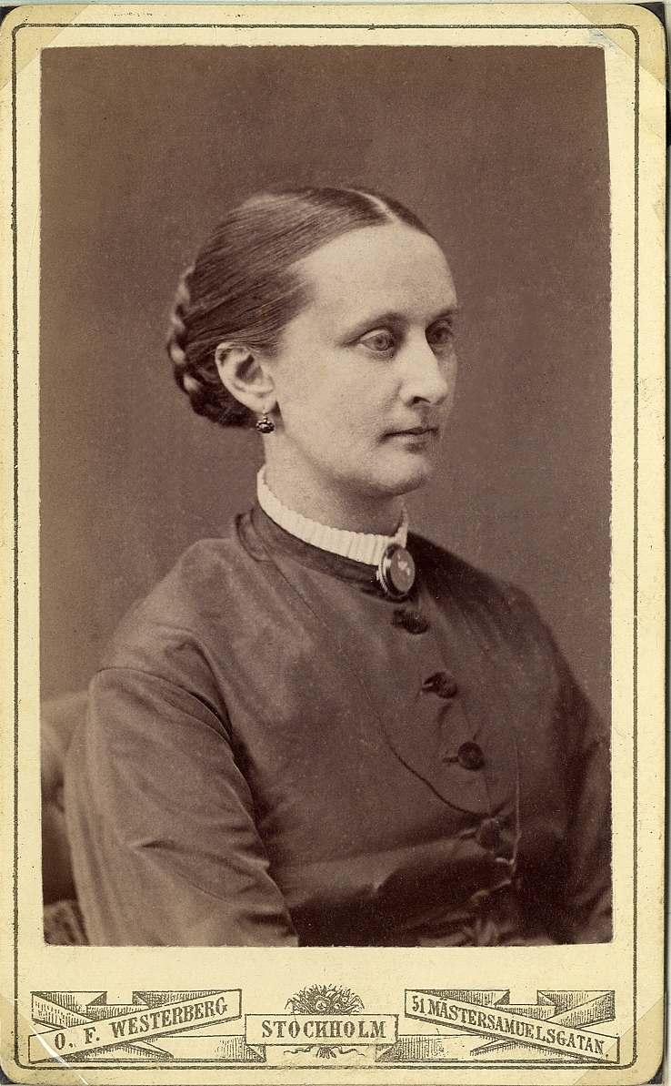 Porträttfoto av en kvinna i mörk sidenklänning med vit ståkrage. Vid kragen syns en brosch och en tunn klockkedja. I öronen syns örhängen. 
Bröstbild, halvprofil. Ateljéfoto.