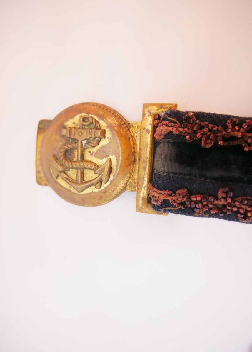 Mørkeblått belte med et svart fløyelsbånd midt på og to rader med brunrøde perler. En metallspenne med ankermotiv. 