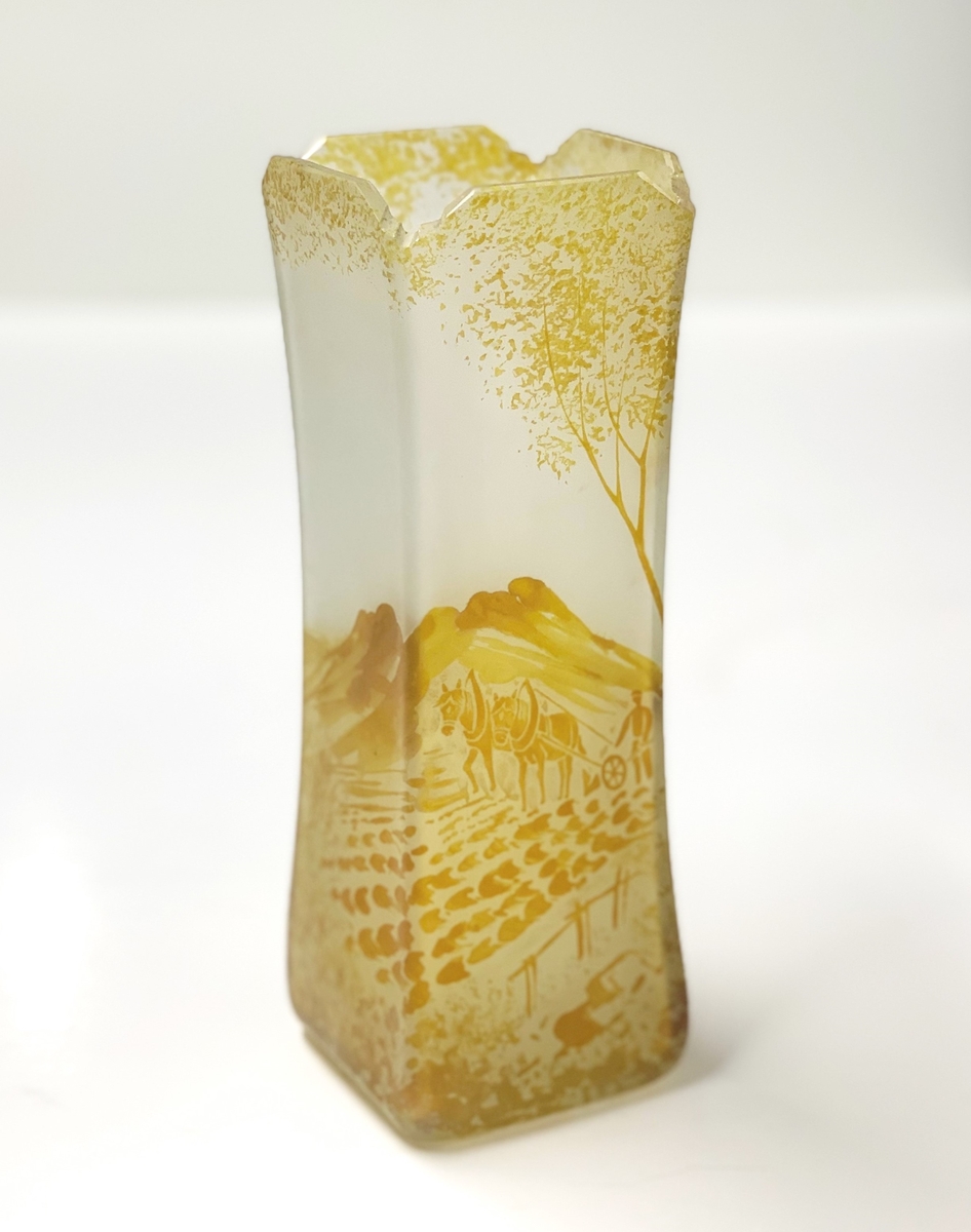 Etsad och målad kvadratisk vas, som smalnar av på mitten. Mynningens fyra hörn är profilslipade och vasens målade motiv består av ett ruralt, bergigt landskap där en man plöjer åkern med ett tvåspann hästar. Vasen bildar parvas med M 14692.
