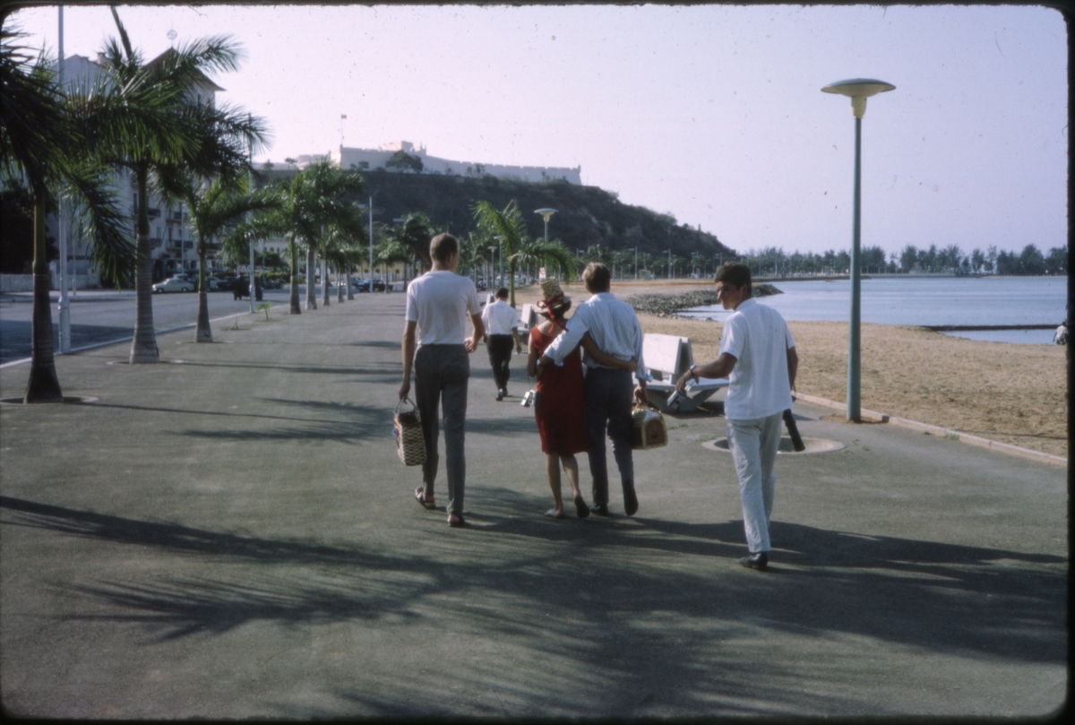 Fem personer går langs en strandpromenade, med en festning i bakgrunnen. 'Sagafjord' Around The World via Africa Cruise 1966.