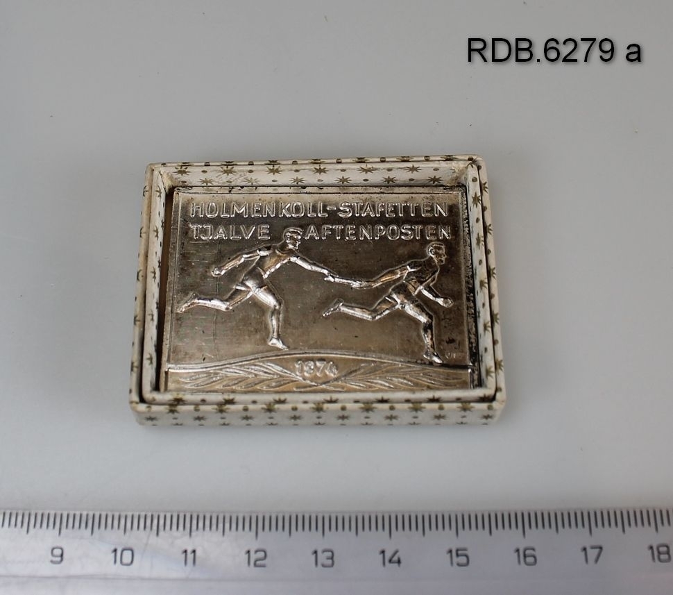 Firkantet liten plakett i sølvfarget metall med relieff av to stafettløpere og innskrift. Plaketten ligger i en hvit eske med små gullstjerner.