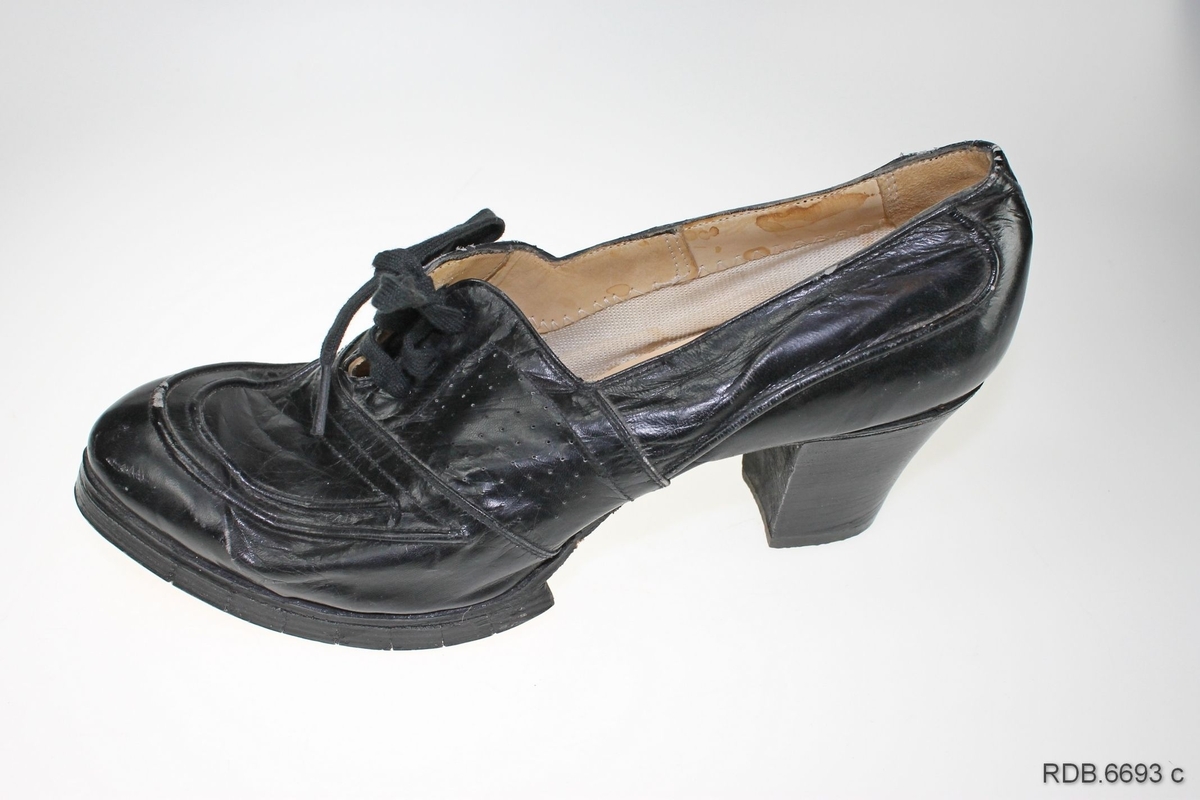 Et par svarte damesko med høge, kraftige hæler. Skoene er "krigsvare", fra 2. verdenskrig, med overlær av tynt stoff og plast og tresåler (trebunner) som er spikret på. Skoene har åpning foran med tunge og tre par hull med itredde svarte lisser. Under hælen er påspikret en gummisåle.