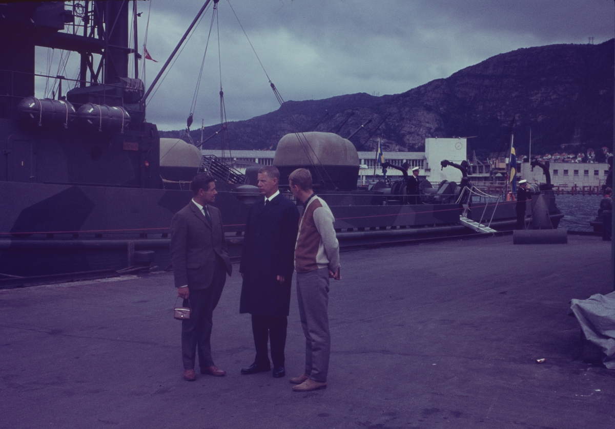 Bilden visar tre män i civila kläder i ett samtal. De står på en kaj med minsveparen Tärnö i bakgrunden.