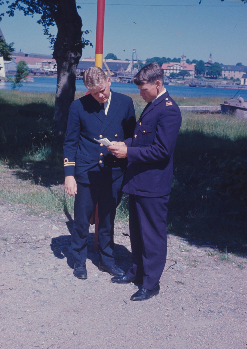 På bilden syns två sjöofficerare i tjänstegrad löjtnant i samtal på Lindholmen. I bakgrunden ser man örlogsvarvet i Karlskrona.