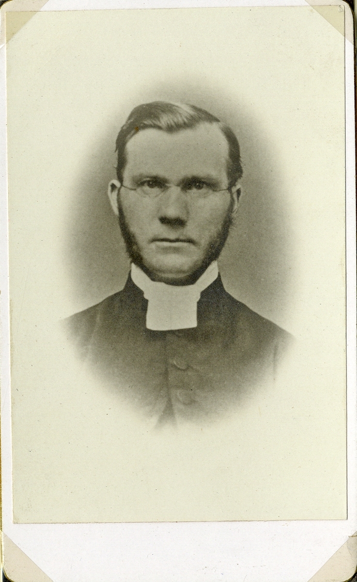 Porträttfoto av en man med glasögon, klädd i prästrock med prästkrage. 
Bröstbild, en face. Ateljéfoto.
