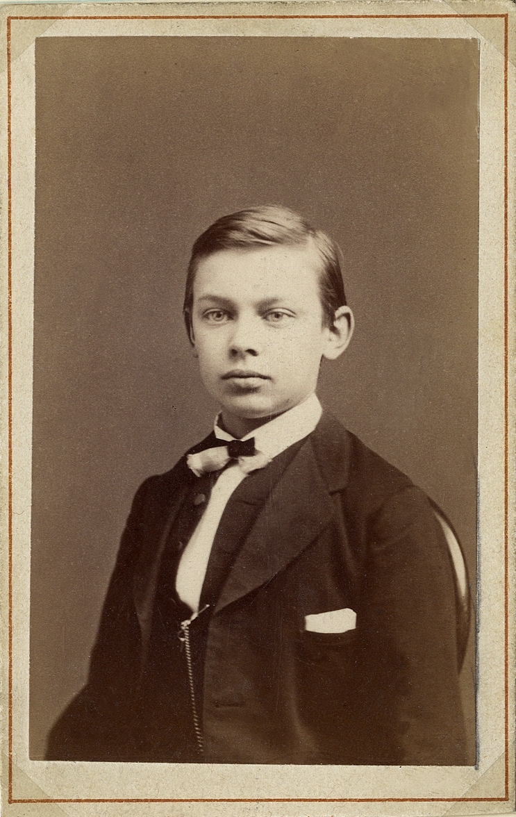 Porträttfoto av en ung man i mörk kavajkostym med väst och fluga. På västen skymtar en klockkedja.
Bröstbild, halvprofil. Ateljéfoto.
