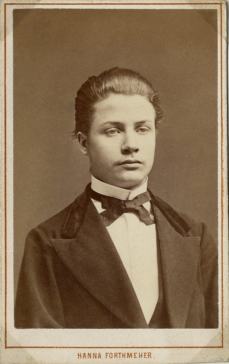 Foto av en ung man i mörk kavajkostym med sammetskrage, väst, stärkkrage och fluga.
Bröstbild, halvprofil. Ateljéfoto.