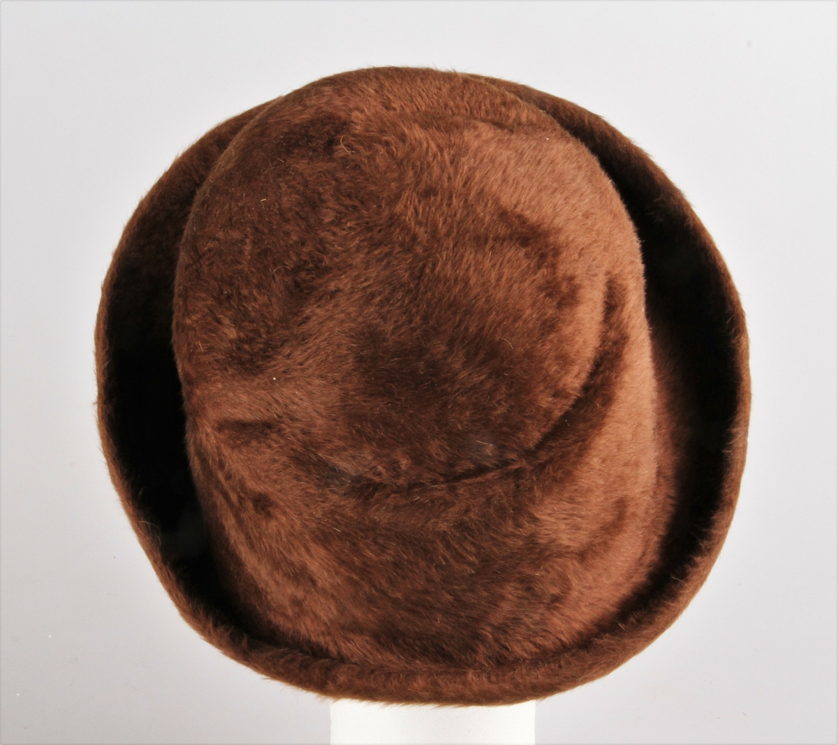 Damehatt laget av hårfilt. Hatten er presset av en grunnform, en cappelin. Pull og brem i ett. Pullen høyere foran og har noen buede linjer som markerer formen. Bremmen er smal bak, bred og oppbrettet foran. Filten er brettet utover og stukket ned med maskin til ca 1 cm bred fald. 1,5 cm bredt, brunt ripsbånd er sydd på ved hodefestet og i båndet er det festet en brun hattestrikk.