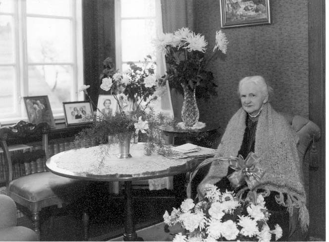 Porträtt av Albertina Kellner i samband med hennes 90-årsdag. En vithårig dam som sitter i en fåtölj i sitt hem med en sjal över axlarna, omgiven av blomsterbuketter.