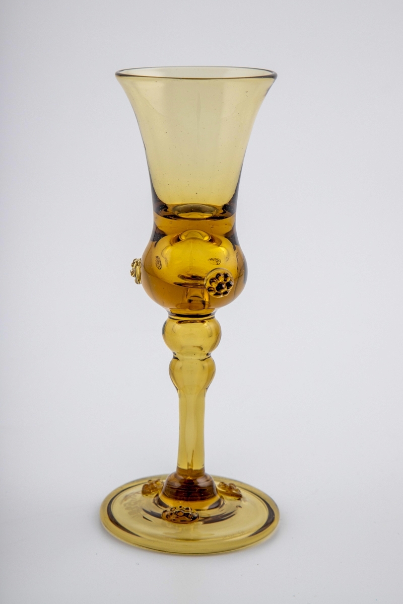 Drammeglass i gjennomskinnelig gulbrunt glass. Øvre del av kupa er konisk, mens nedre del er kuleformet og dekorert med tre pålagte rosettligende ornamenter ("bringebær"). Høy stett med to vulster. Foten er svakt buet med forhøyet kant, dekorert med tre rosettligende ornamenter.