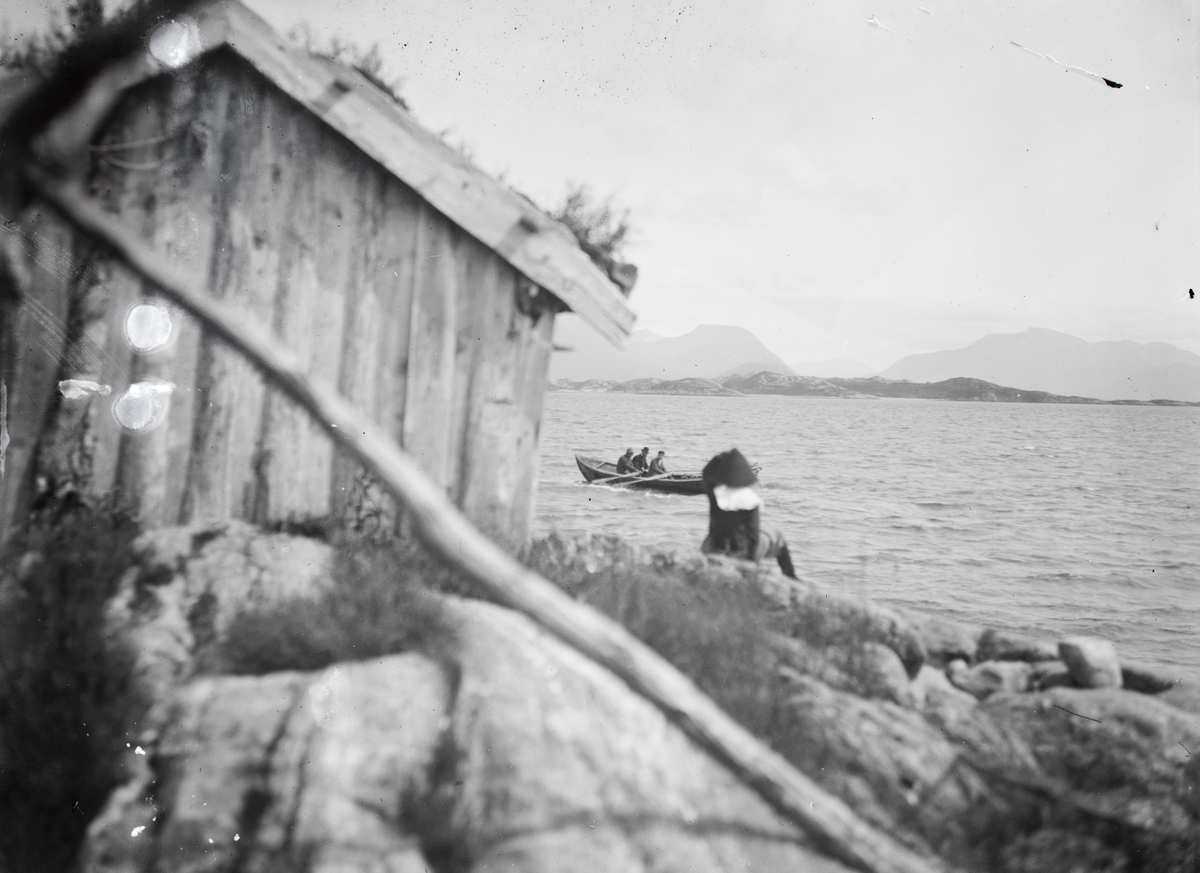 Motiv av en robåt på fjorden. I forgrunnen sitter en gutt ved et naust.
