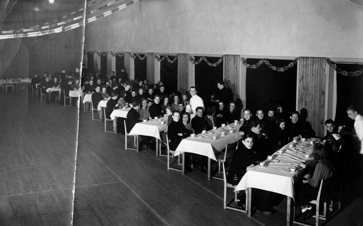 Festsammankomst på F 7, tidigt 1940-tal. Män i uniform och kvinnor sitter vid bord i en gympasal. Kaffe serveras.